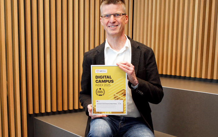 Ein Mann sitzt vor einer Wand aus Holzlamellen auf einer Treppe und präsentiert die gerahmte Urkunde 
