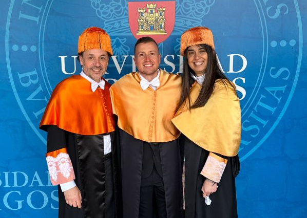 Zwei Männer und eine Frau Arm in Arm in Doktorroben vor dem Wappen der Universidad de Burgos