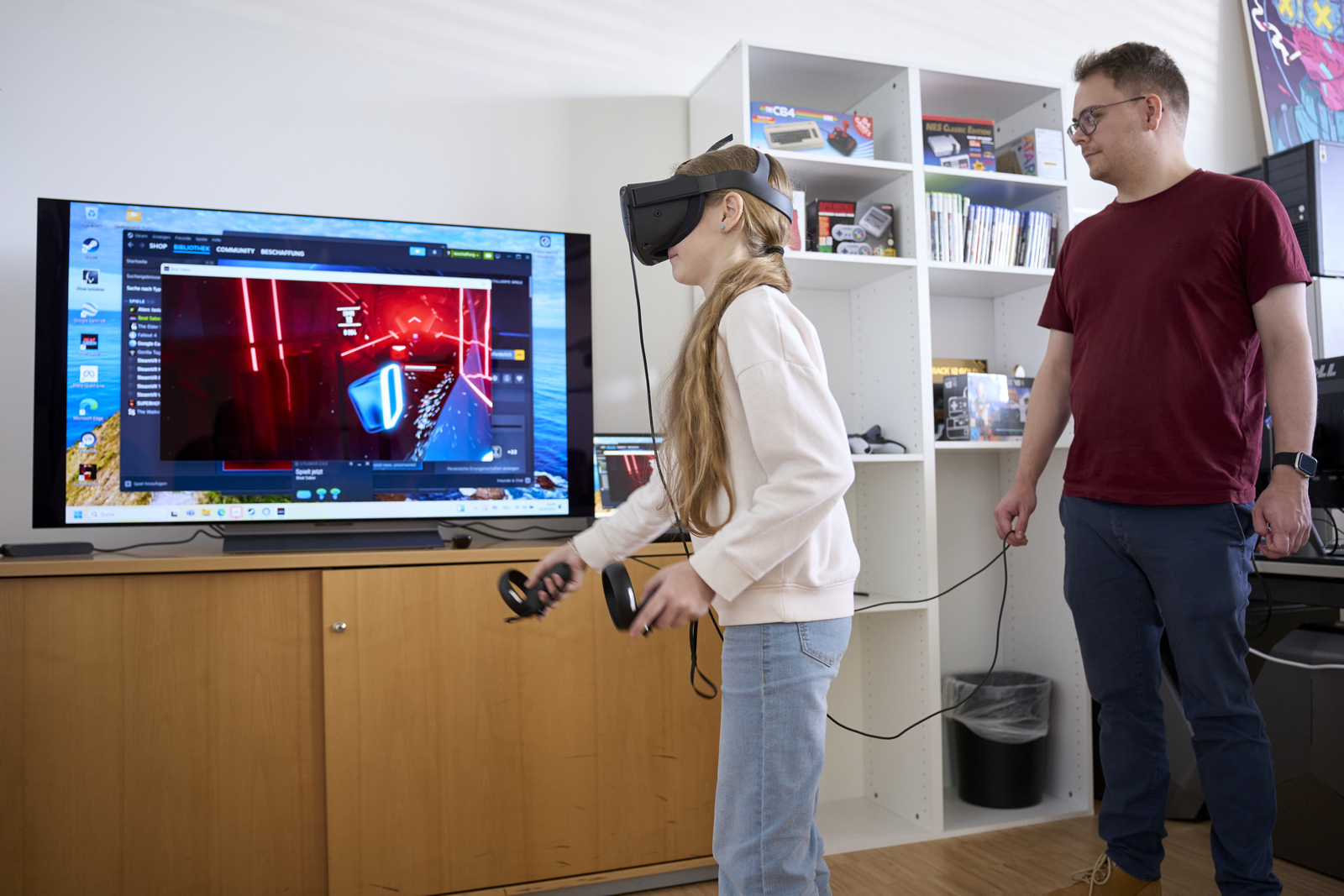 Ein Mädchen mit einer VR-Brille spielt ein Spiel auf einem Bildschirm, ein junger Mann beobachtet sie