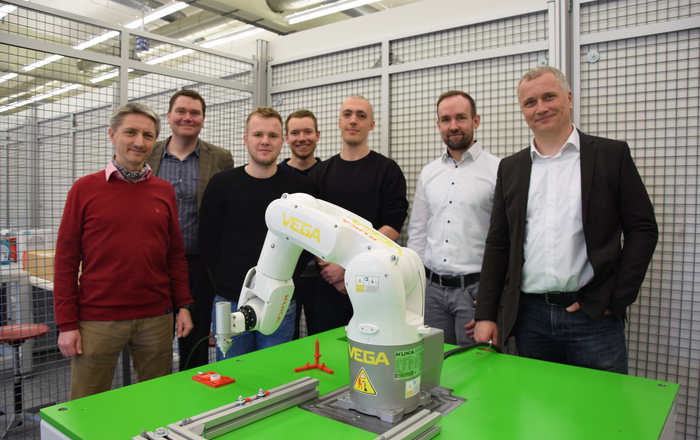 Die Projektbeteiligten stehen hinter dem Tisch mit dem Roboter der programmiert wird.