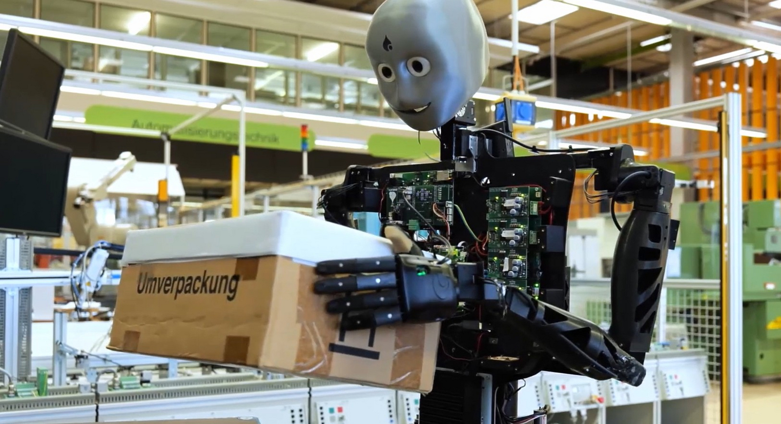 Ein humanoider Roboter steht vor einer Maschine und hält ein Paket vor einen Bildschirm.  