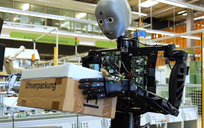 Ein humanoider Roboter steht vor einer Maschine und hält ein Paket vor einen Bildschirm.  
