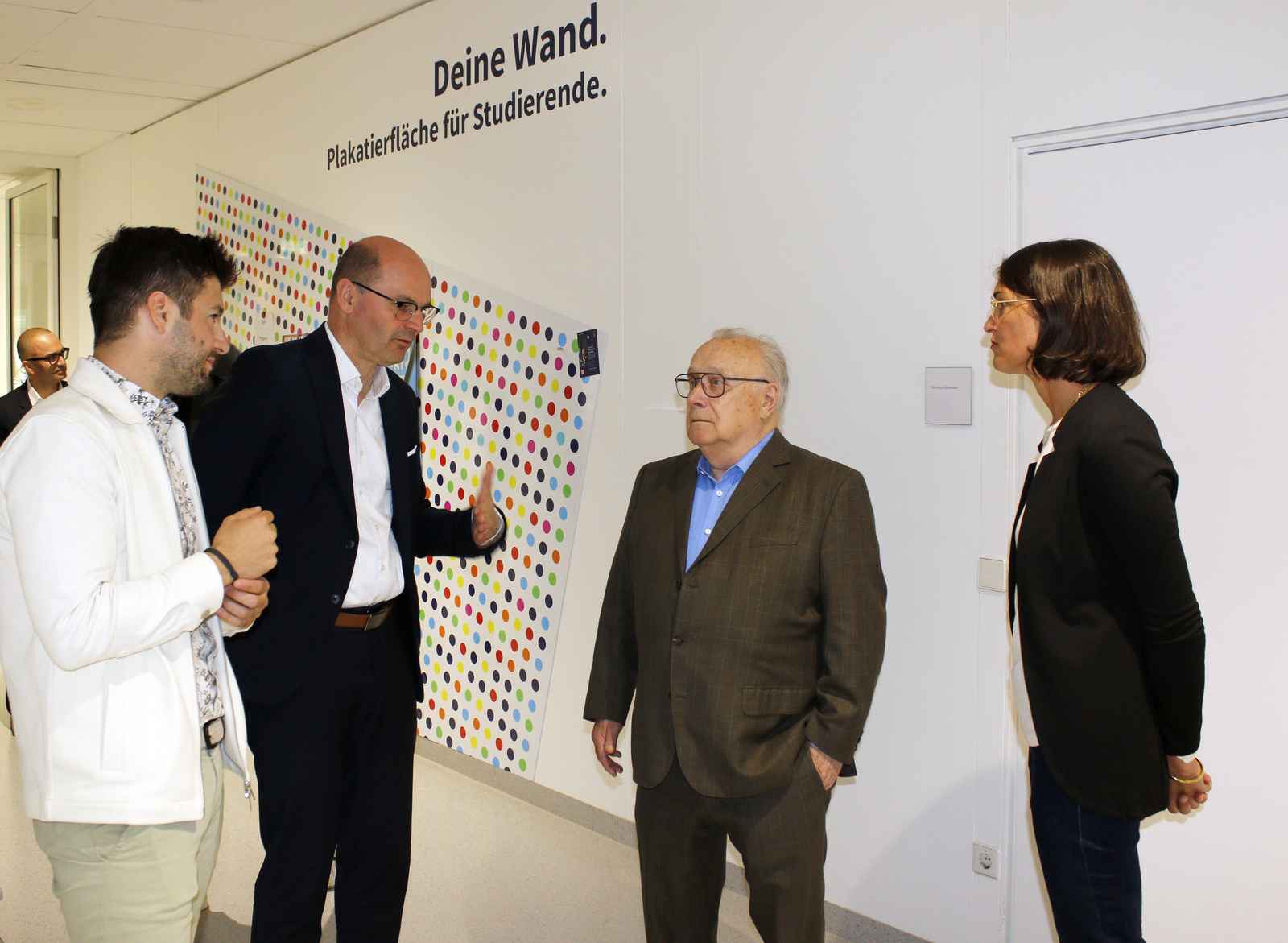 Vier Personen stehen vor einer grafisch designten Pinwand und unterhalten sich