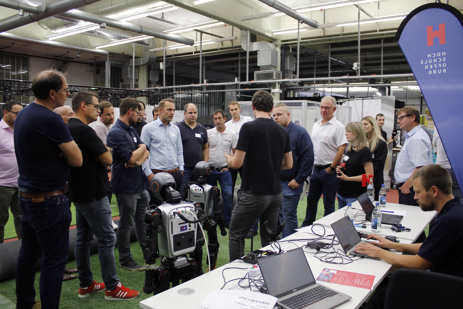 Ganz viele Menschen stehen um zwei humanoide Roboter herum, ein Mann erklärt etwas