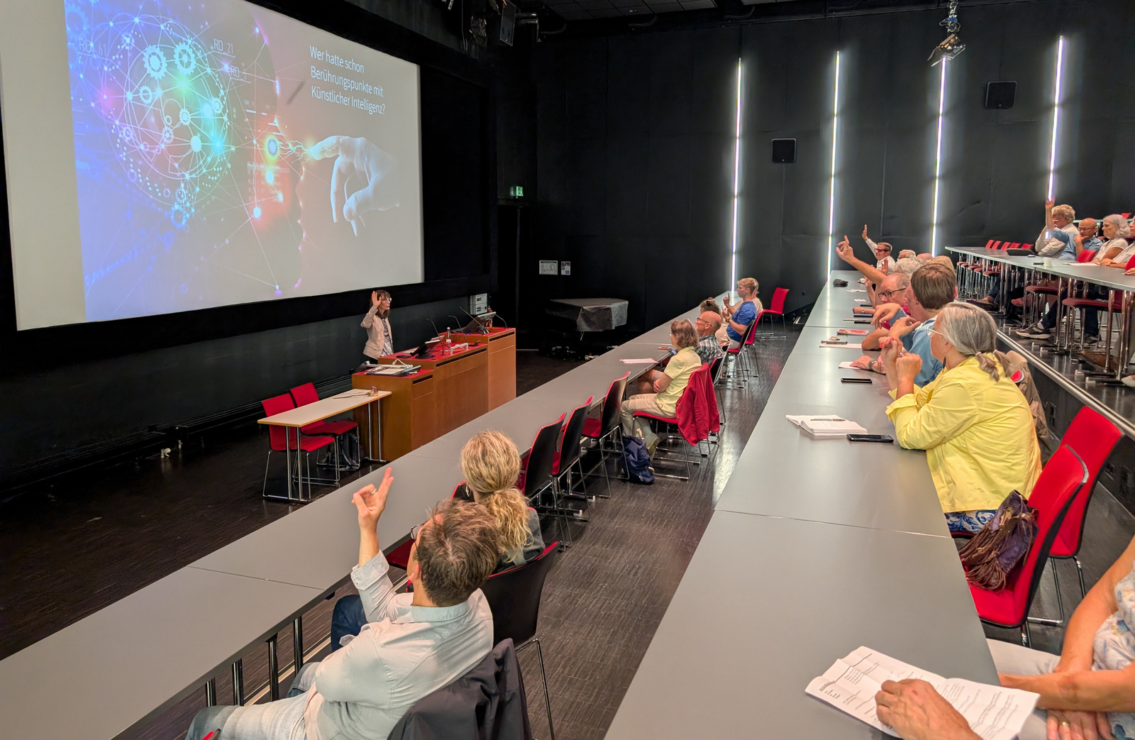 Eine Frau steht vor einer großen Leinwand und spricht zu sitzenden Zuhörenden in einem Hörsaal