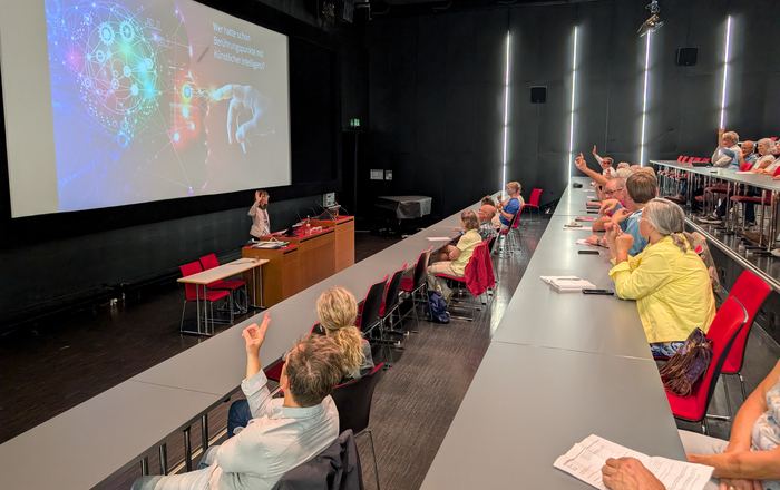 Eine Frau steht vor einer großen Leinwand und spricht zu sitzenden Zuhörenden in einem Hörsaal