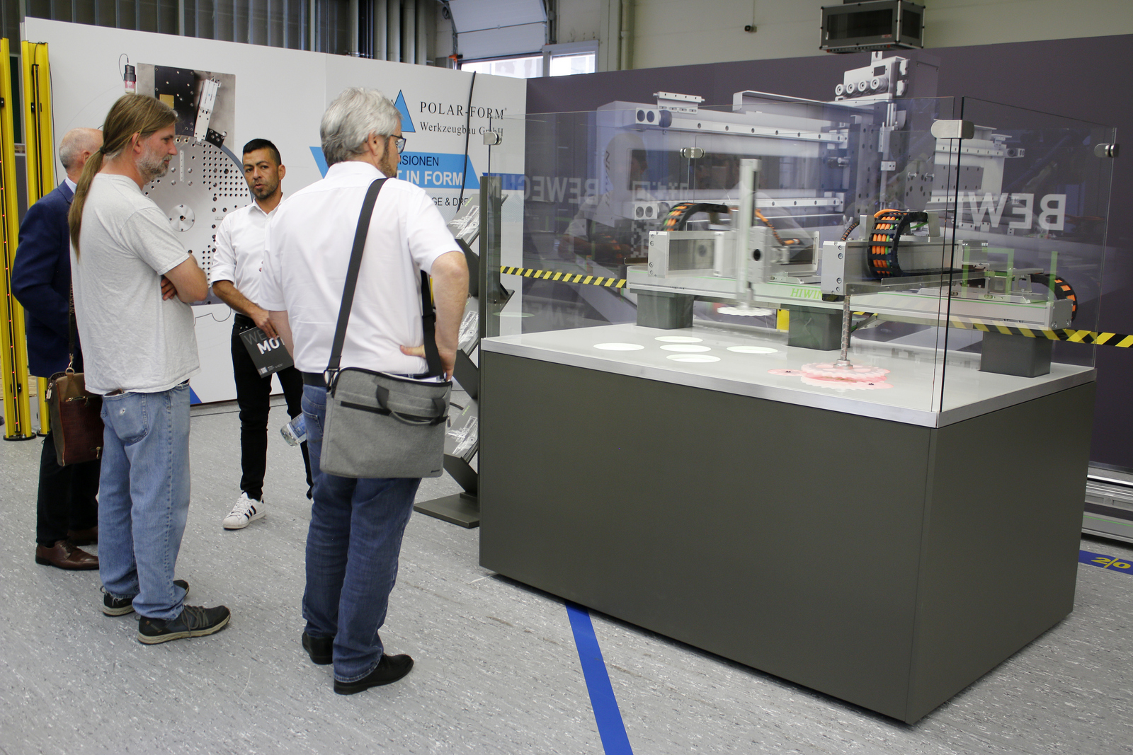 Vier Männer stehen um eine Glaskasten mit dem Modell einer Linearachse herum, einer erklärt etwas