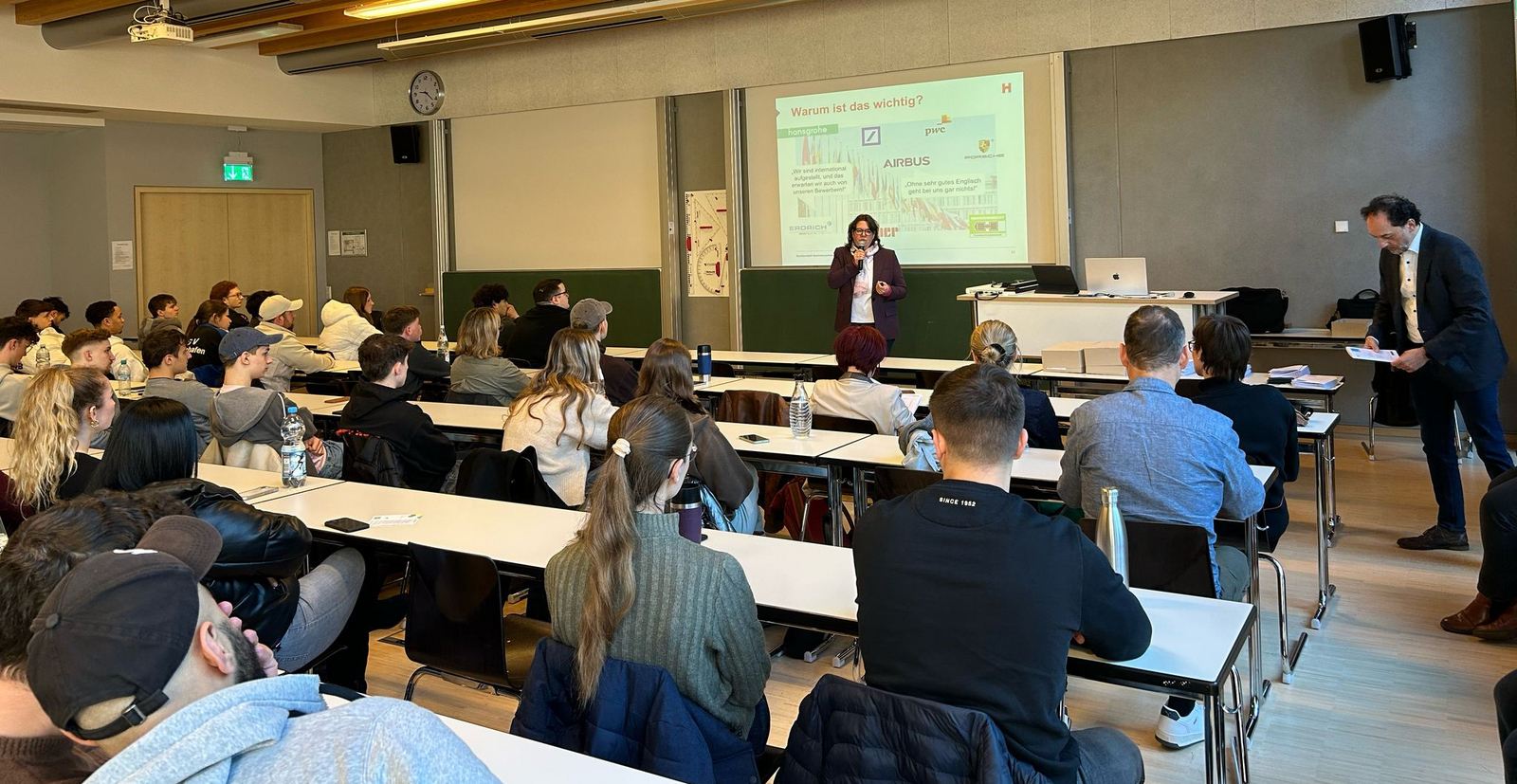 Eine Frau steht vor an Tischen sitzenden jungen Leuten vor einem Bildschirm