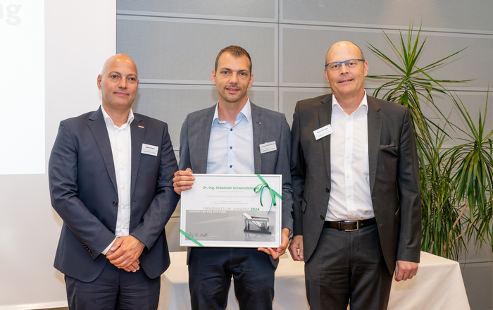 Drei Männer stehen nebeneinander, der Mittlere hat eine gerahmte Urkunde in der Hand