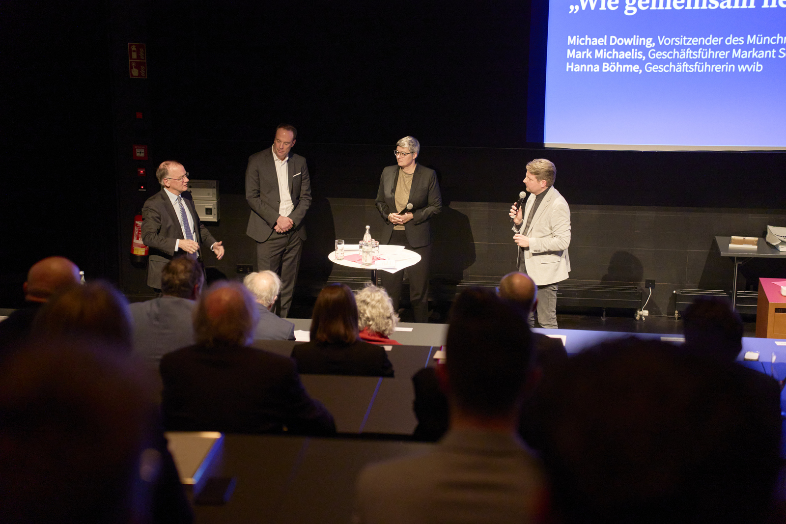Drei Männer und eine Frau stehen mit Mikrofonen im Halbkreis um einen Stehtisch herum und sprechen miteinander