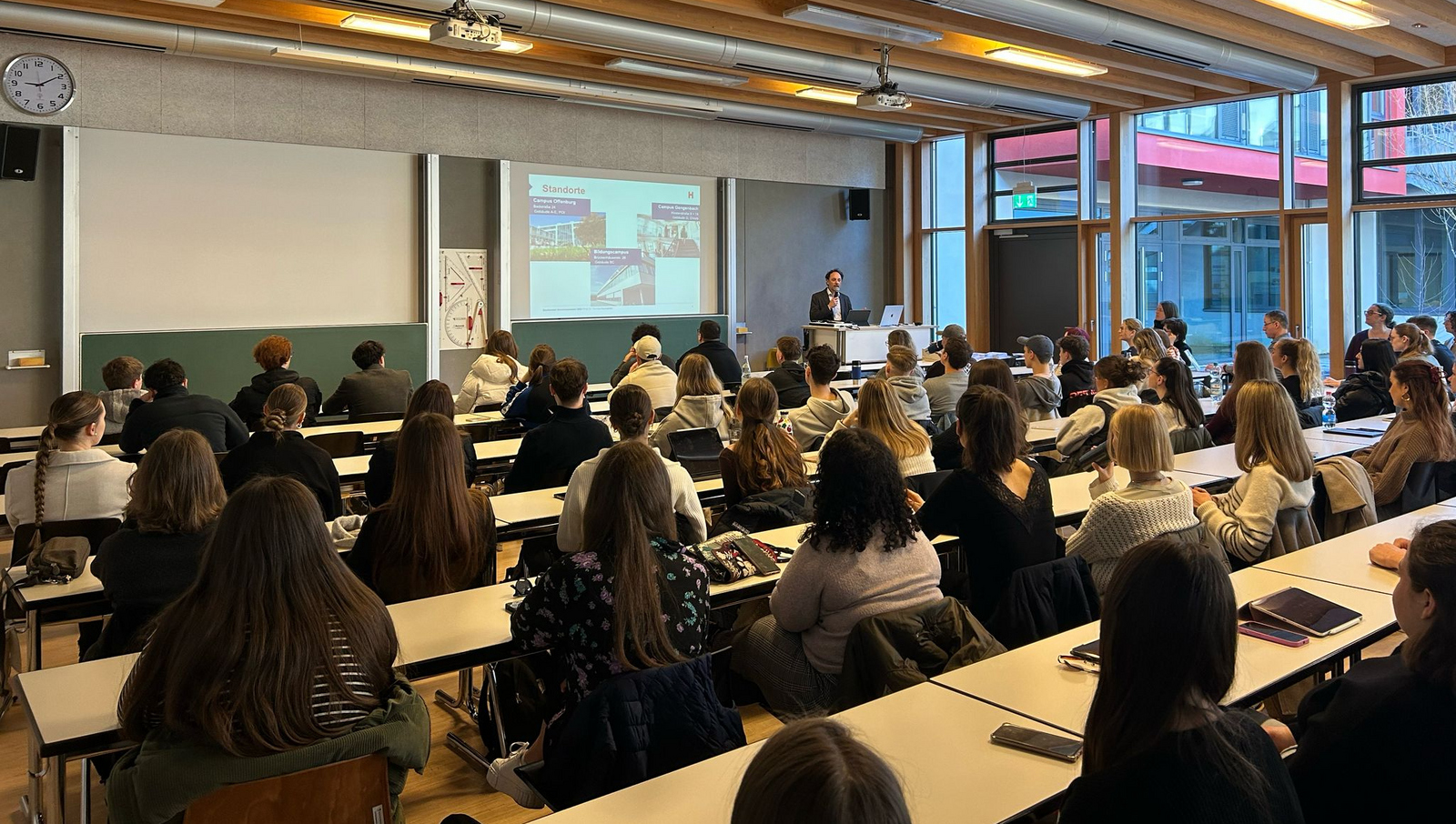Ein Mann steht an einem Rednerpult vor an Tischen sitzenden jungen Leuten und erläutert eine Präsentation auf einem Bildschirm