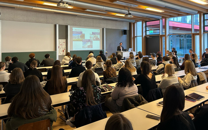 Ein Mann steht an einem Rednerpult vor an Tischen sitzenden jungen Leuten und erläutert eine Präsentation auf einem Bildschirm