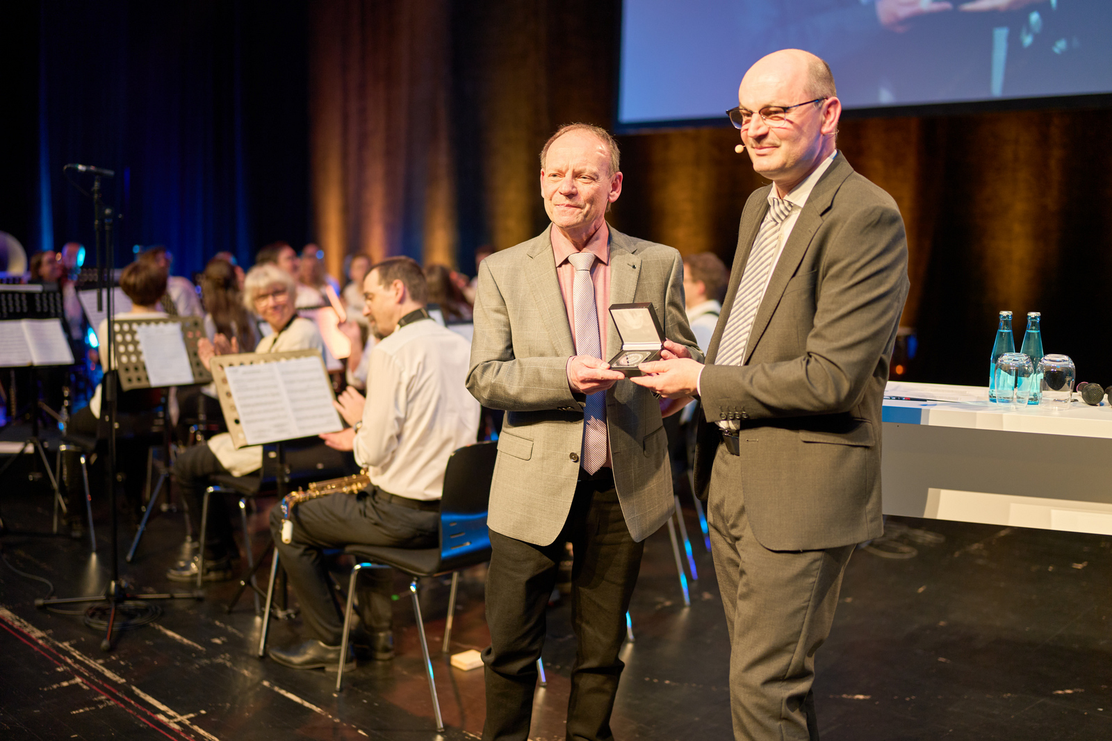 Zwei Männer im Anzug stehen vor einem sitzenden Orchester, der Rechte überreicht dem Linken eine Ehrenmedaille