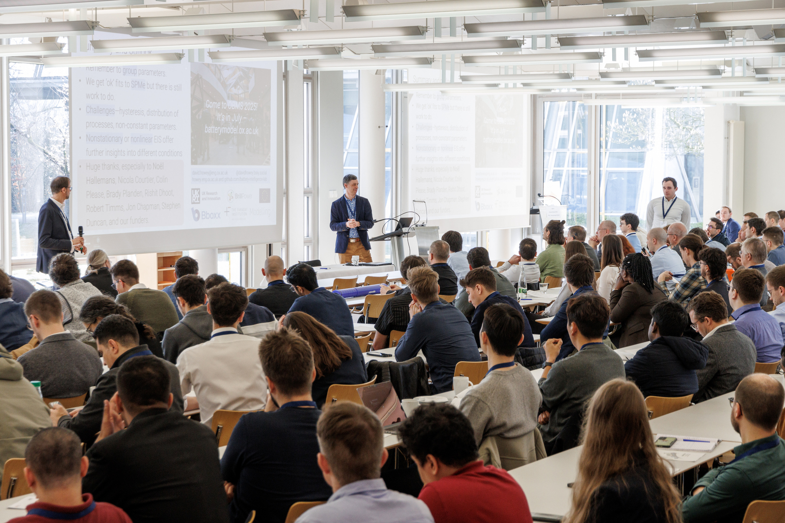 Zwei Männer stehen vor zahlreichen sitzenden Konferenzteilnehmenden