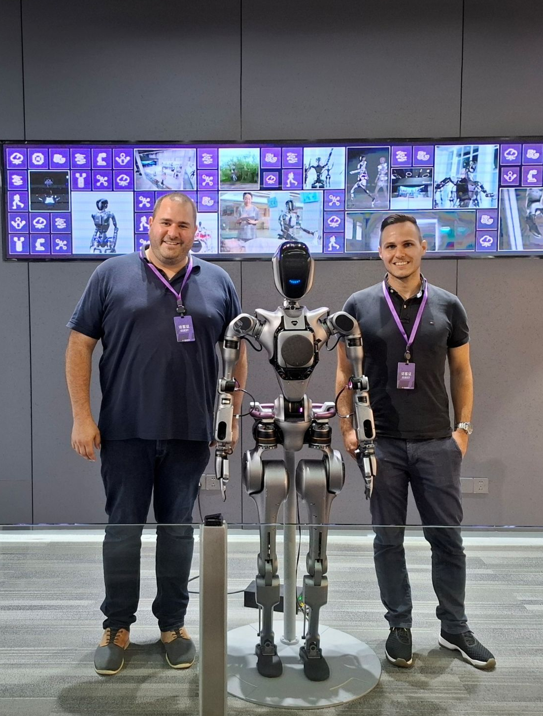 Zwei junge Männer stehen rechts und links von einem humanoiden Roboter, im Hintergrund sind Bildschirme zu sehen