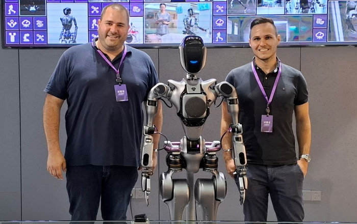 Zwei junge Männer stehen rechts und links von einem humanoiden Roboter, im Hintergrund sind Bildschirme zu sehen