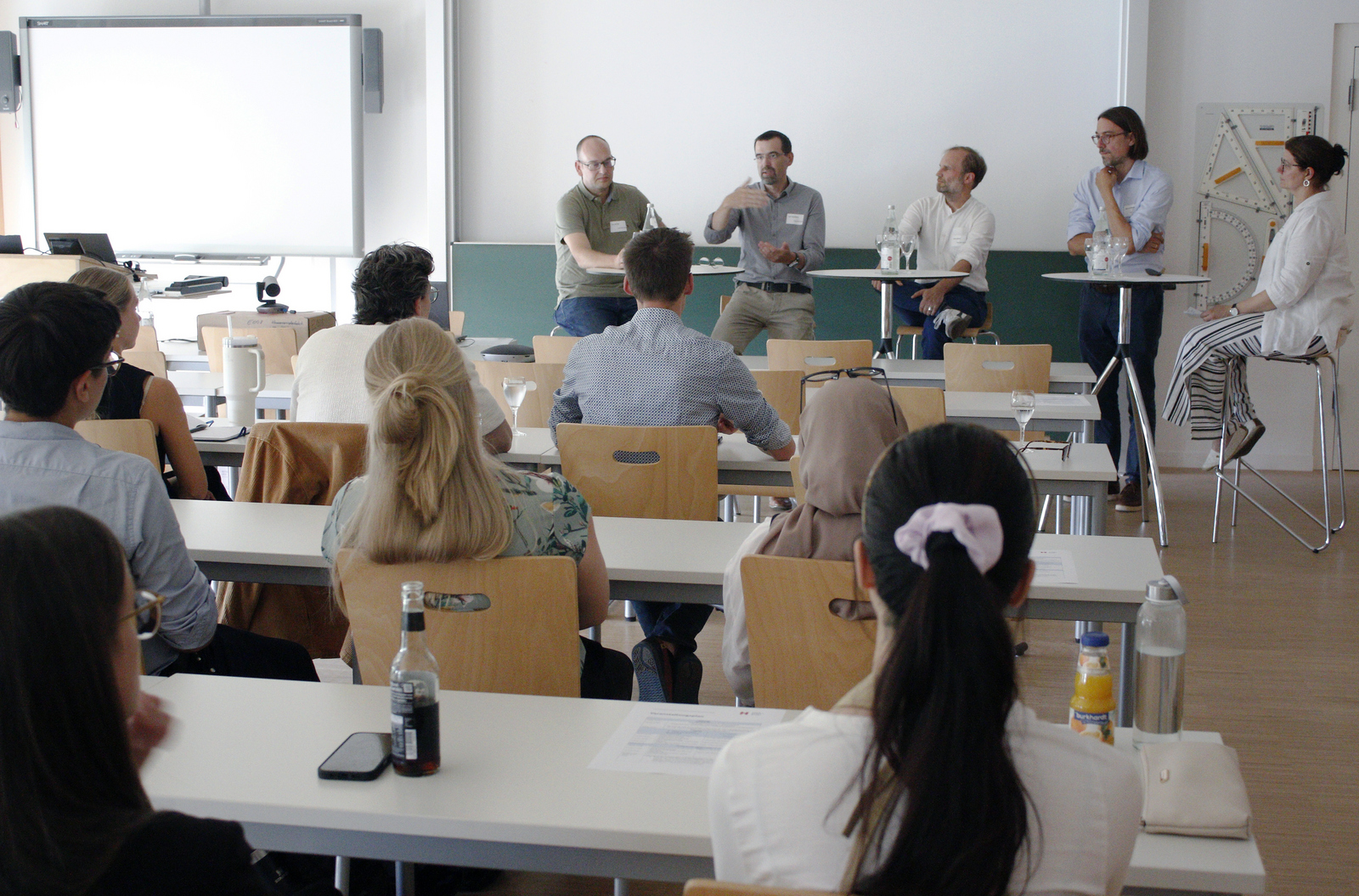 Vier Männer und eine Frau sitzen auf Barhockern an Stehtischen und diskutieren