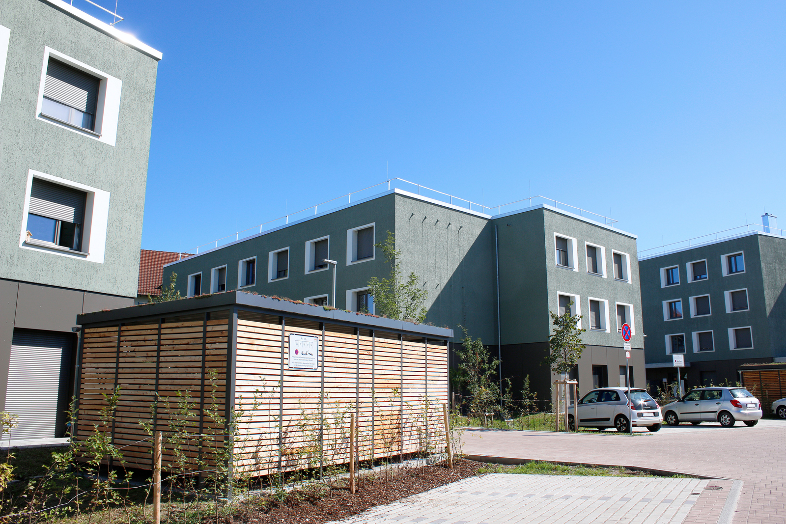 Drei grün-braune Gebäude, davor Parkplätze und ein Fahrradunterstand aus Holz 