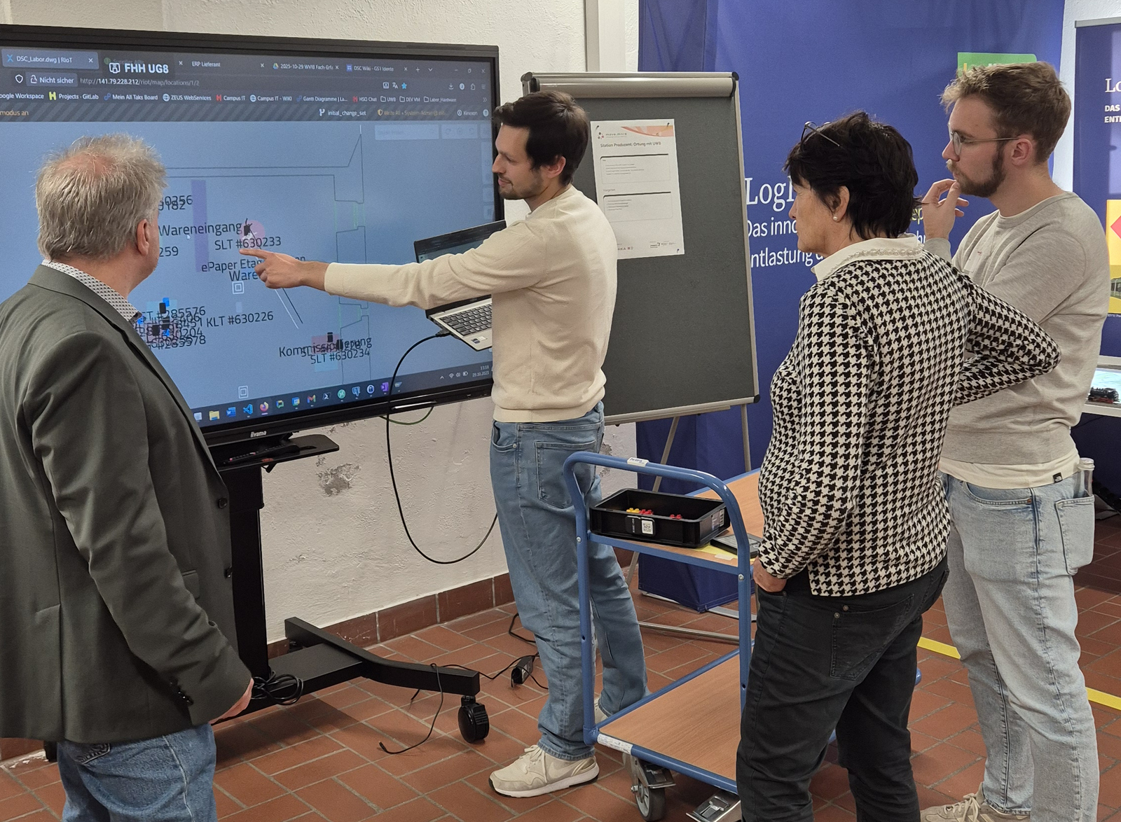 Vier Personen stehen vor einem großen Bildschirm mit Diagrammen, eine Person zeigt darauf.