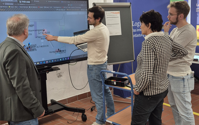 Vier Personen stehen vor einem großen Bildschirm mit Diagrammen, eine Person zeigt darauf.
