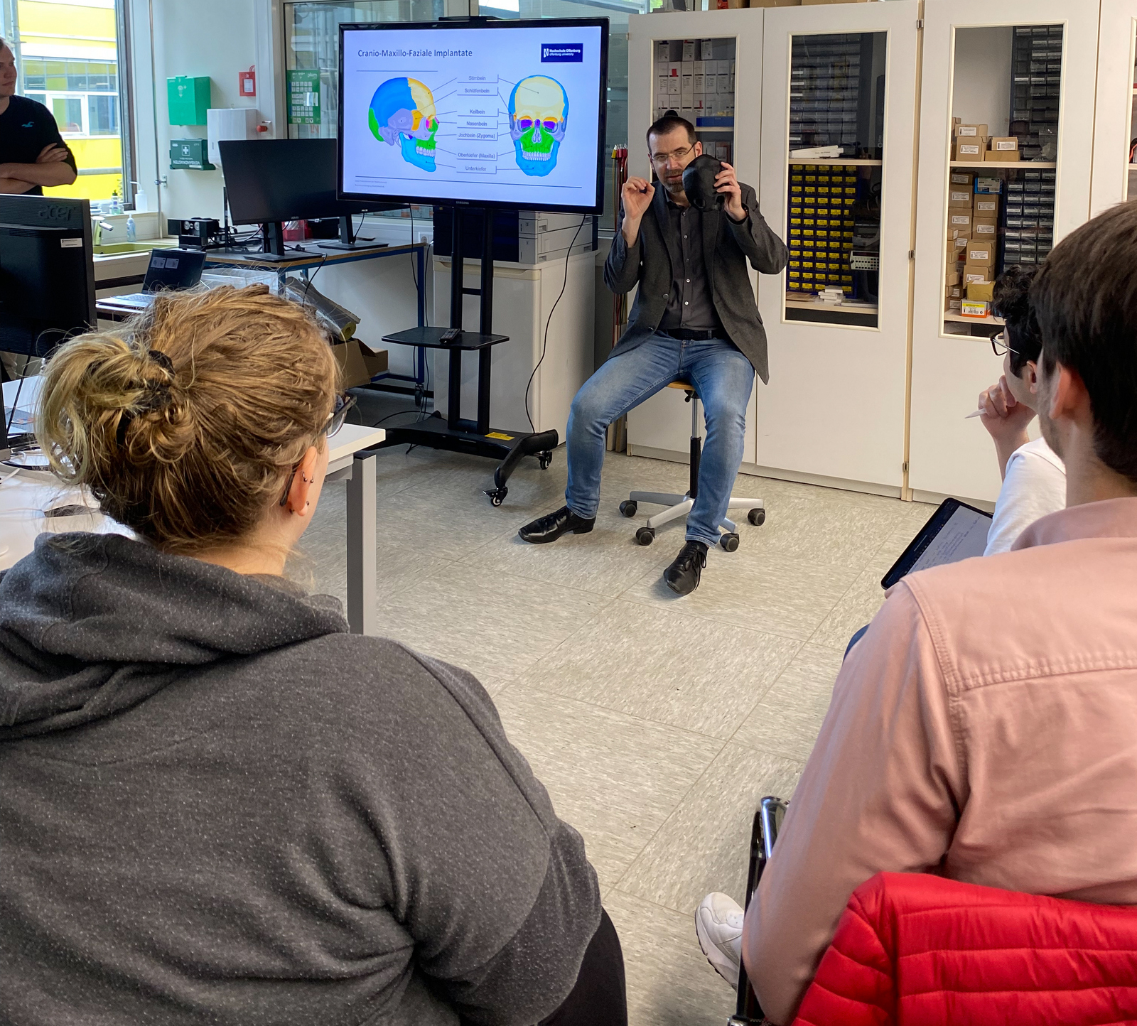 Prof. Peter Quadbeck sitzt vor einigen Studierenden und neben Bildschirm auf dem eine Skizze den Schädelaufbau zeigt