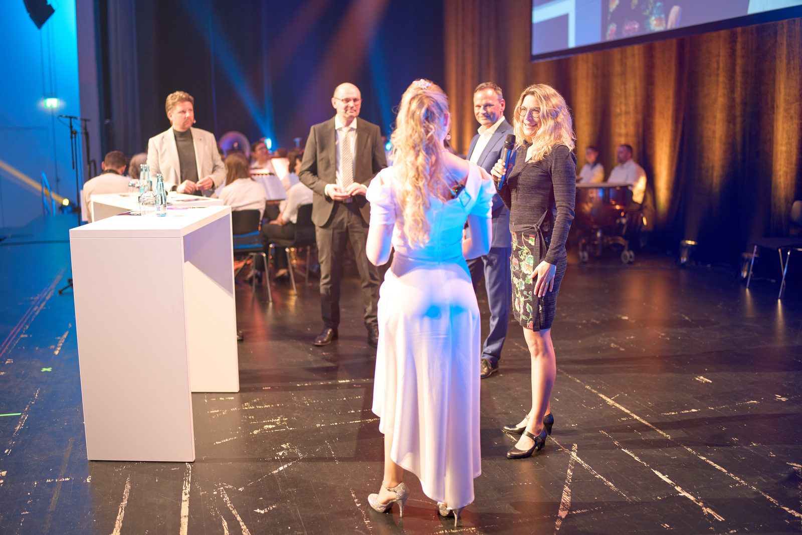 Drei Männer und zwei Frauen stehen auf der Bühne im Halbkreis um eine Tisch und unterhalten sich