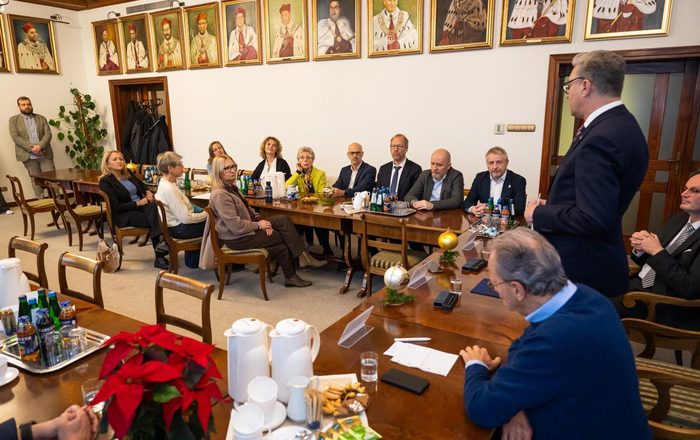 Eine Gruppe Menschen sitzt in eine Saal an dessen Wänden gemalte Porträts von Menschen in rot-weißen Roben hängen