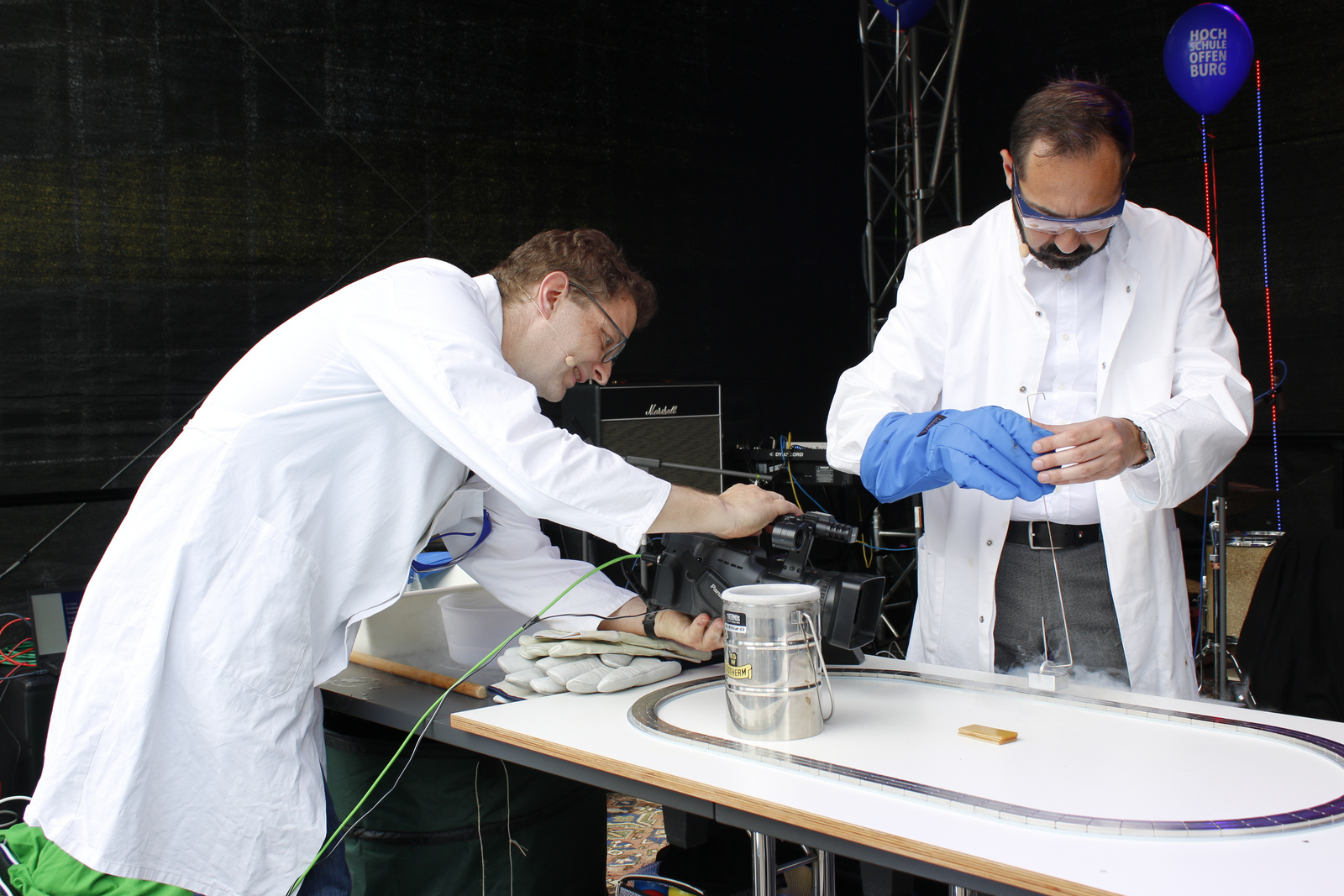 Zwei Männer in weißen Kitteln und mit Schutzbrillen, der eine filmt mit einer Videokamera, der andere lässt einen Magneten schweben. 