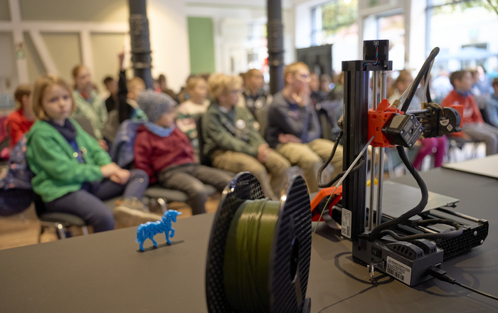 Im Vordergrund ein kleiner 3D-Drucker mit Filament und einem kleinen blauen 3D-Druck eines Pferdes, im Hintergrund die kleinen Besucher der KinderUni