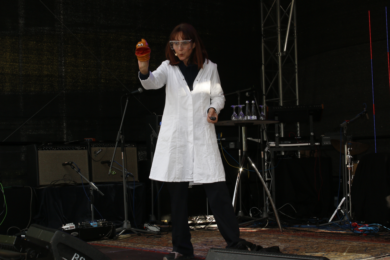 Eine Frau im weißen Kittel mit Schutzbrille und eine Handpuppe in Form eines Kothaufens mit Gesicht