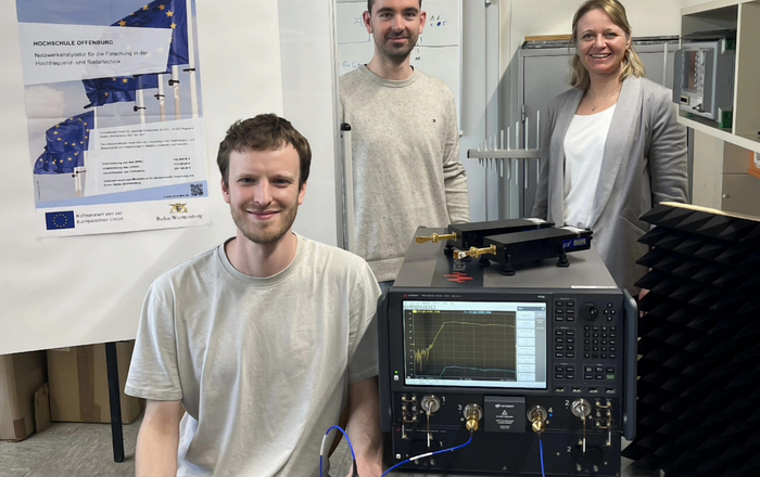 zwei Männer und eine Frau sitzen beziehunsgweise stehen um den Netzwerkanalysator, der auf einem Tisch steht, herum