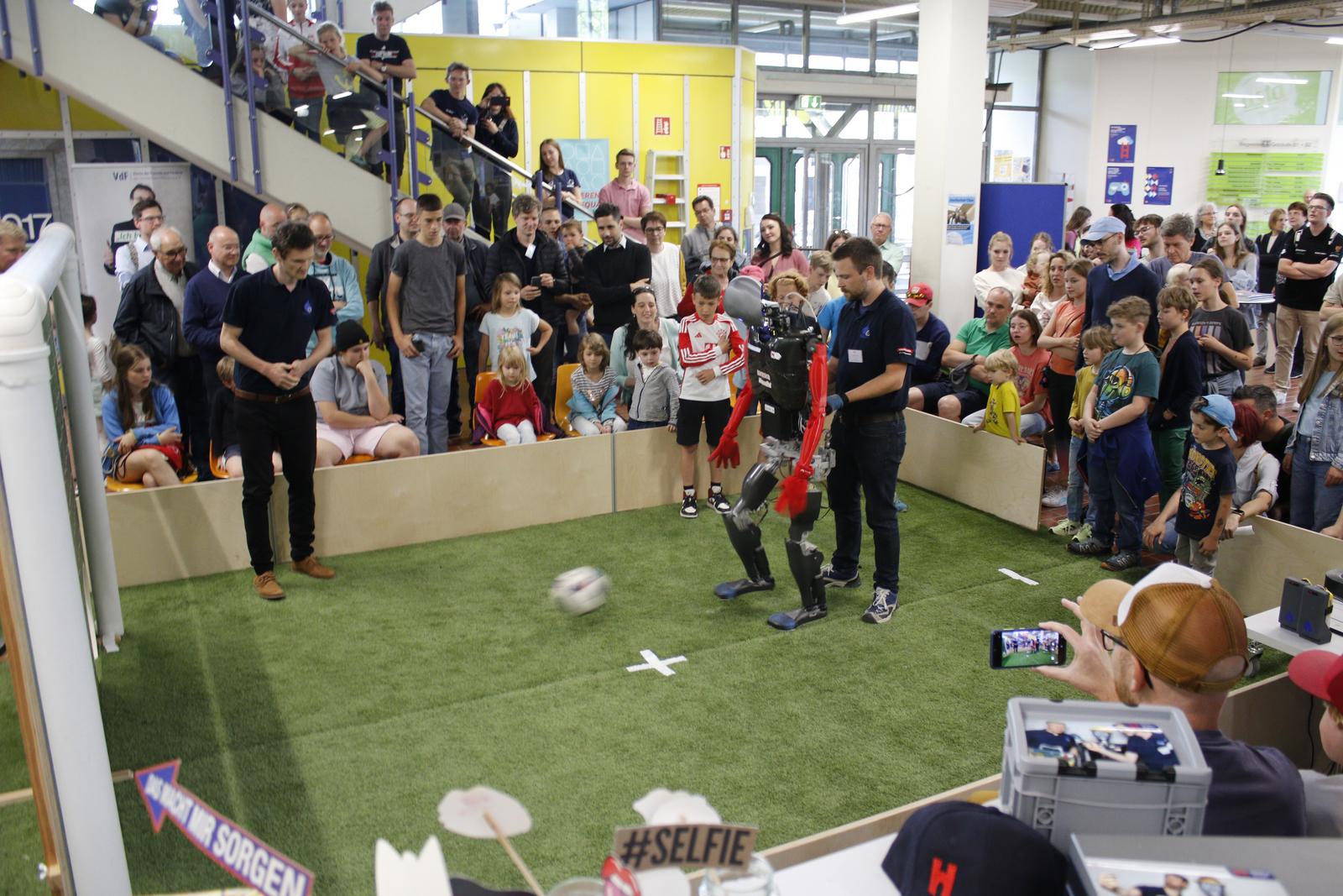 Ein Roboter schießt einen Fußball über das Spielfeld Richtung Torwand während viele andere drumherum stehen.