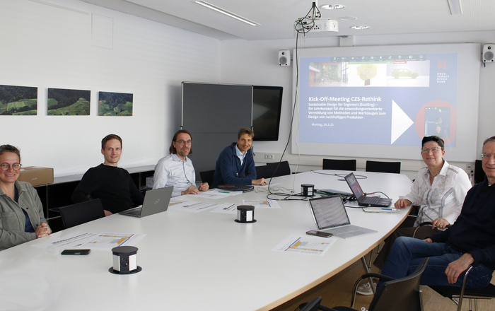 Menschen sitzen um eine Tisch, im Hintergrund ist ein Präsentationsbildschirm