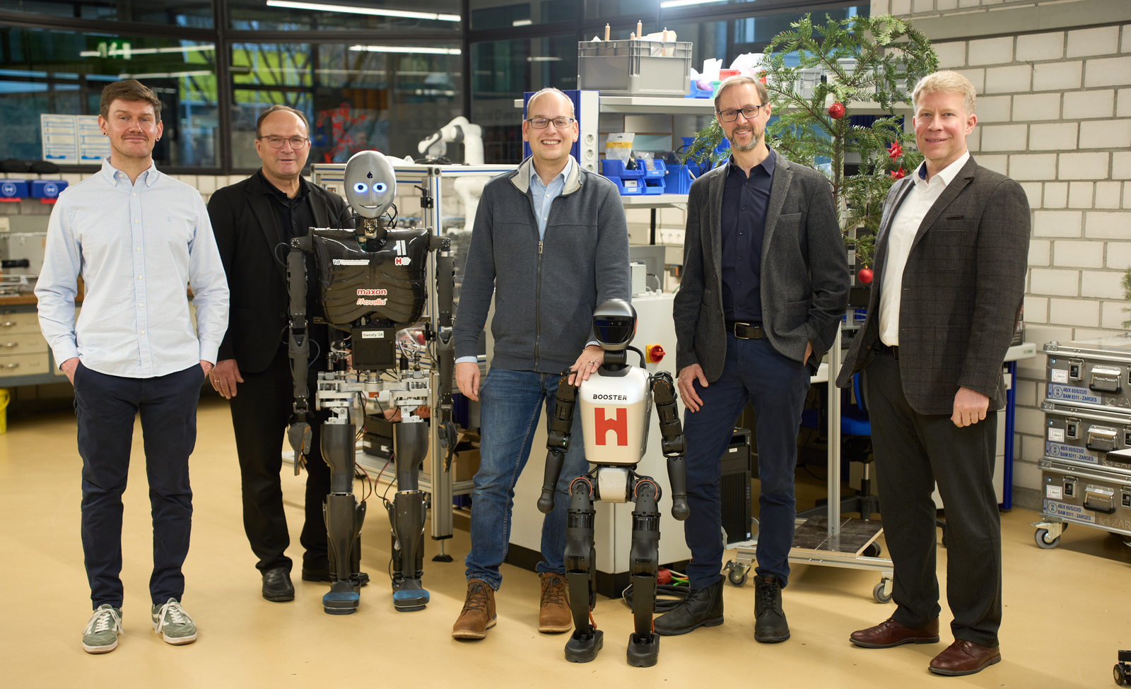 Fünf Männer stehen mit zwei humanoiden Robotern in einem weihnachtlich geschmückten Labor