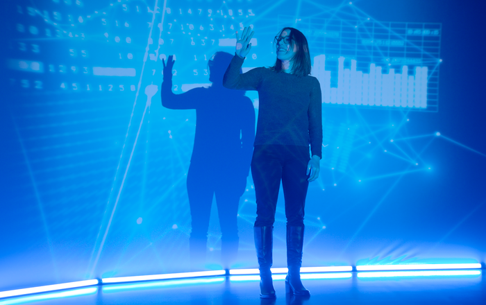 Eine junge Frau steht vor einem Bluescreen auf dem KI-Formeln zu erkennen sind und fährt mit der Hand darüber.