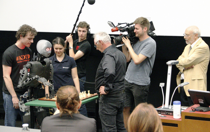 Sweaty und Michael Maly stehen sich an einem Tisch mit einem Schachbrett gegenüber umringt von Studierenden, Kameraleuten, Prof. Dr. Ulrich Hochberg und Zuschauenden