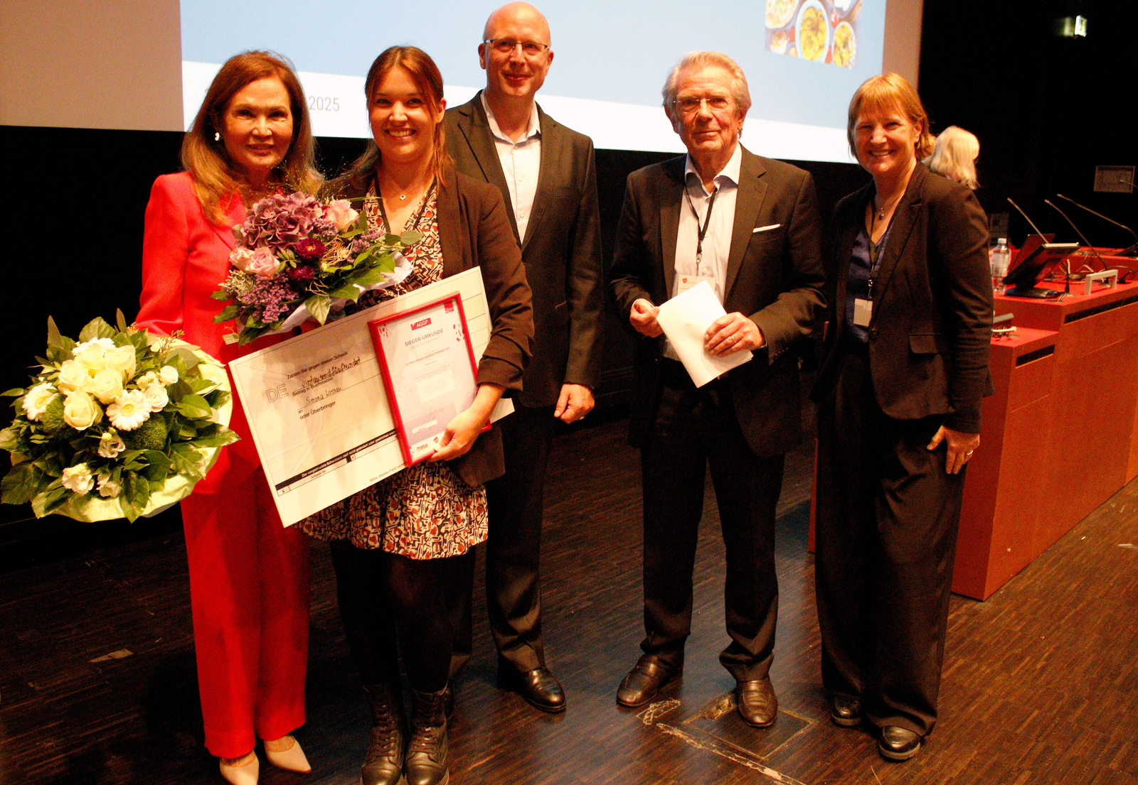 EinE gruppe von chic gekleideten Menschen zwei Frauen haben Blumensträuße in den Händen, die eine zusätzlich noch eine Urkunde und einen Scheck 