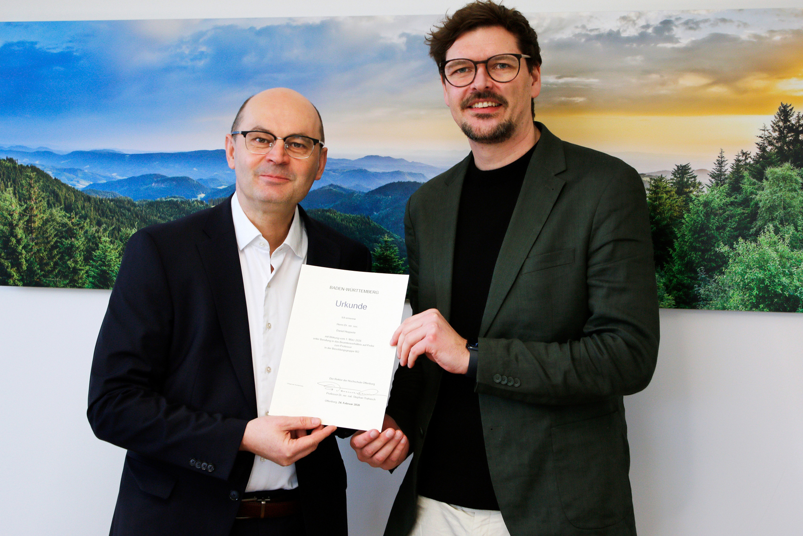 Zwei Männer in Anzügen stehen vor einem Schwarzwaldfoto und halten eine Urkunde zwischen sich