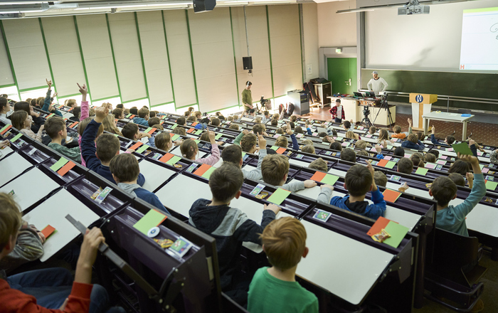 Kinder, die während einer Vorlesung im Saal sitzen. Viele davon melden sich.