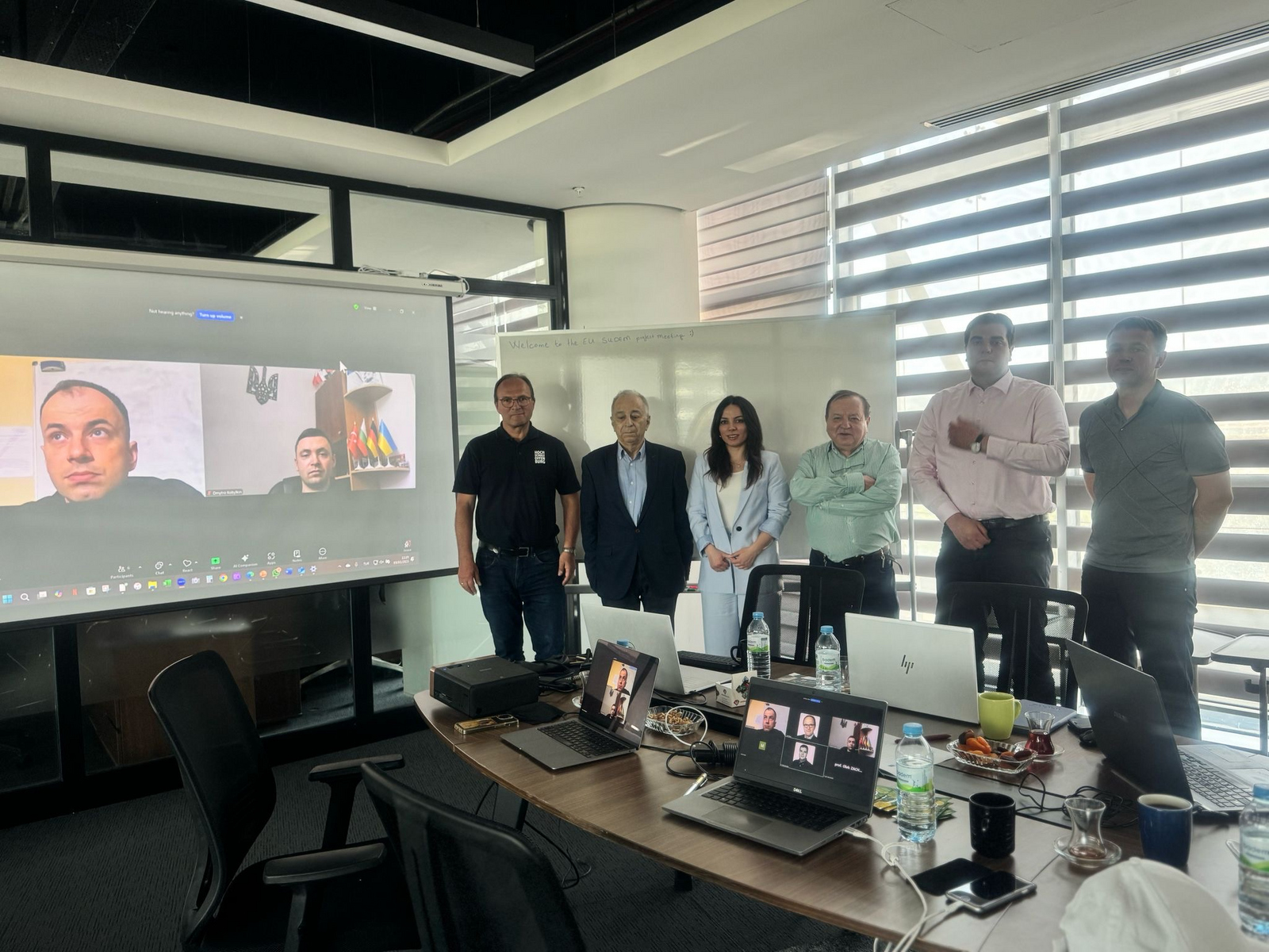 6 Personen in Reihe neben einer Leinwand mit 2 zugeschalteten Personen