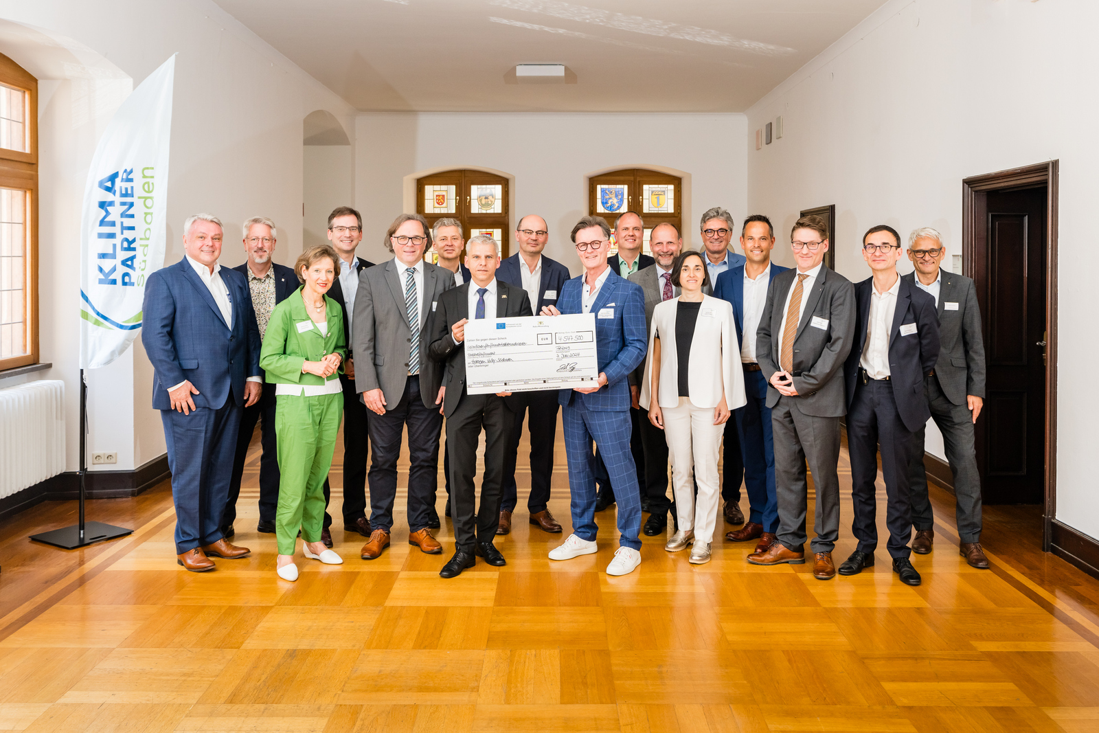 Gruppenfoto mit Vetreterinnen und Vertretern des Konsortiums, Staatssekretär Dr. Patrick Rapp und einem symbolischen Scheck und 