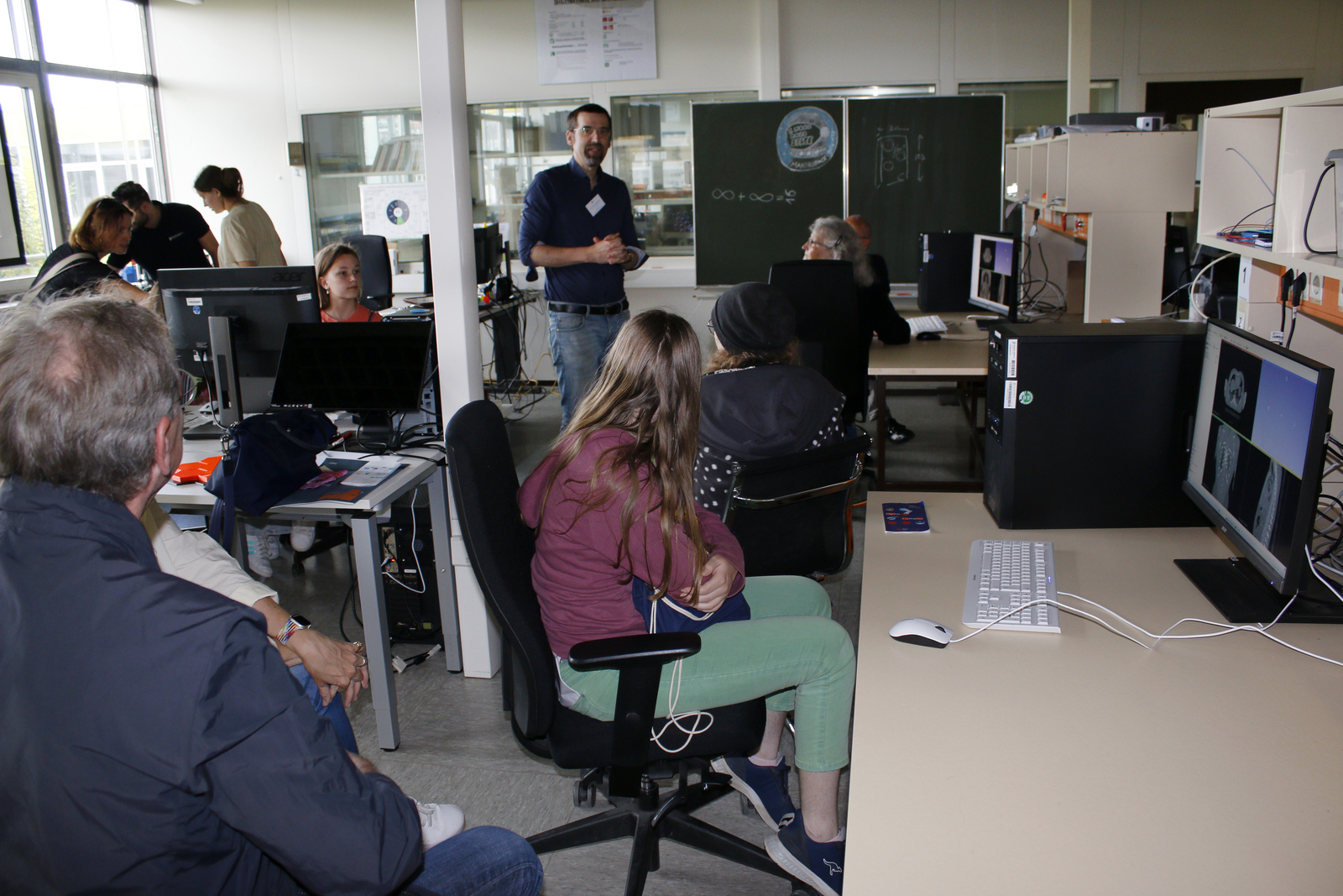 Ein Mann steht im Labor zwischen Besucherinnen und Besuchern, die an Rechnern sitzen oder stehen.