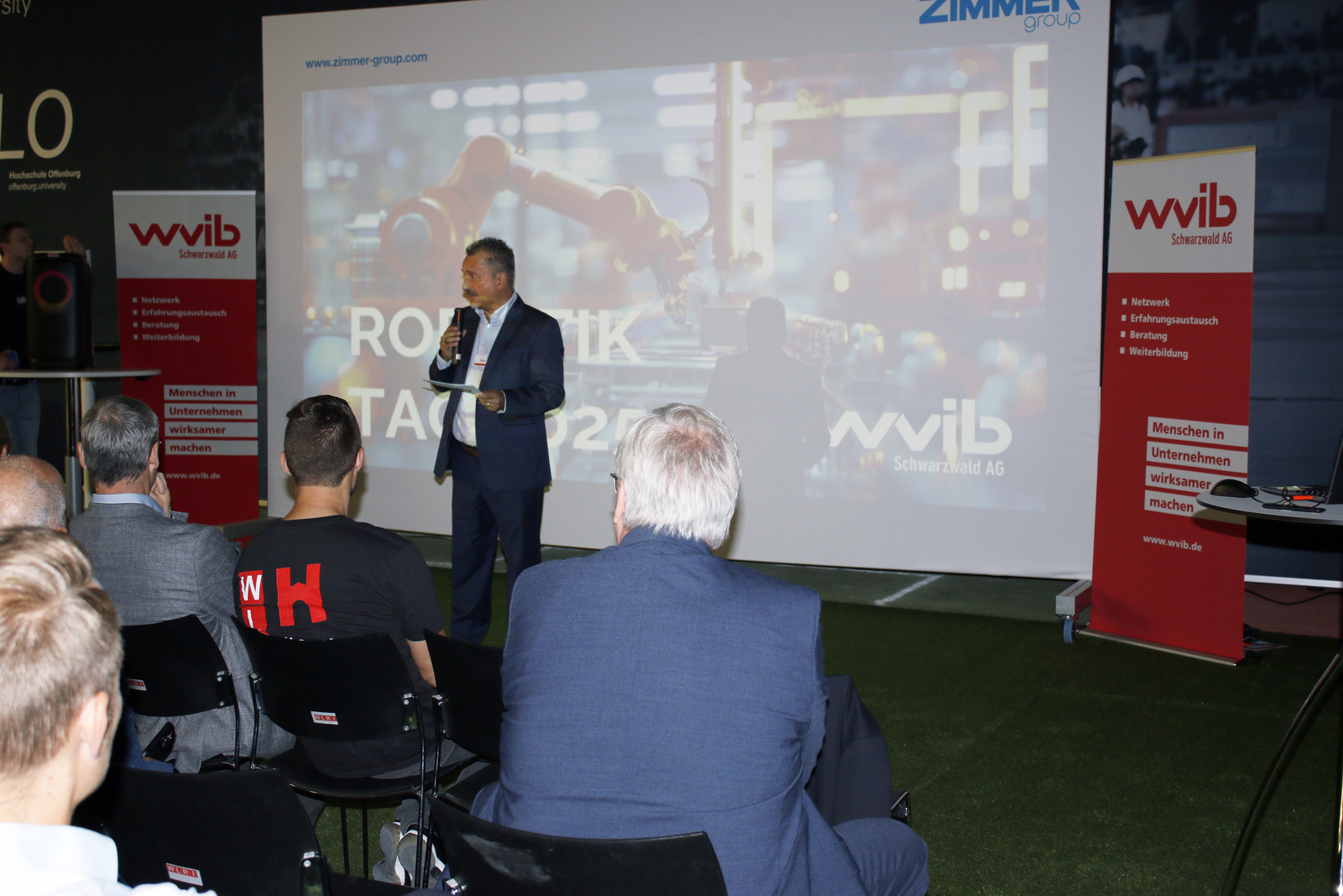 Ein Mann steht vor einer Leinwand vor sitzenden Zuhörenden und spricht ins Mikrofon