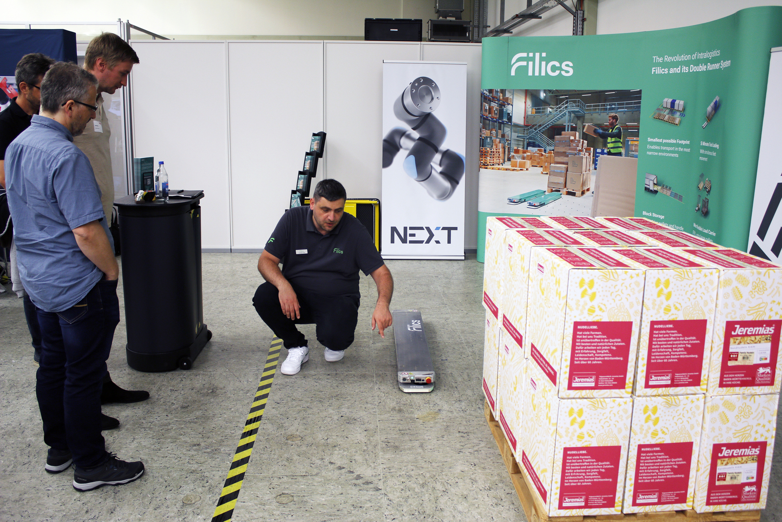 Ein Mann kniet neben einer fahrenden Kufe neben eine Palette und erklärt den Zuschauenden etwas