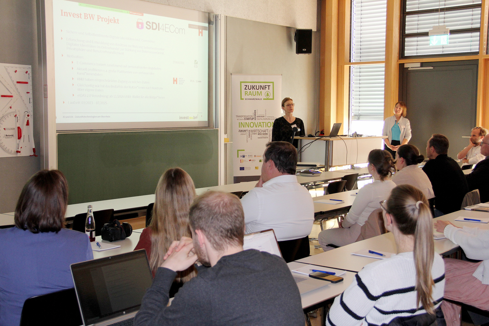 Zwei Frauen stehen rechts einer Leinwand vor dem sitzenden Publikum und erläutern eine Präsentation