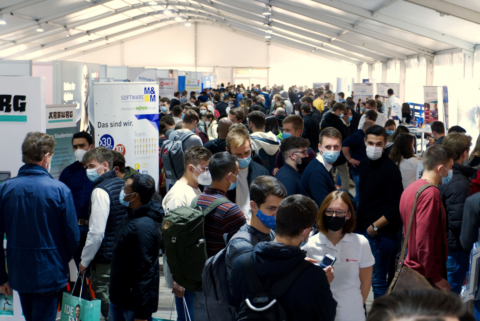 Ausstellende und Studierende mit Maske im Messezelt.