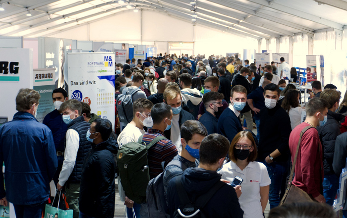 Ausstellende und Studierende mit Maske im Messezelt.