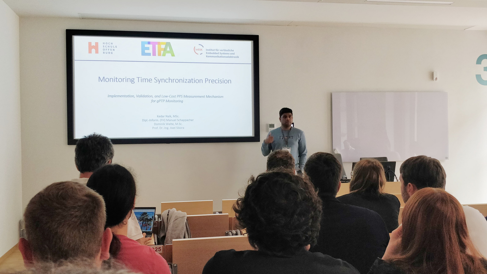 Raum mit Zuhörern 1 Person steht vor einer Leinwand und präsentiert