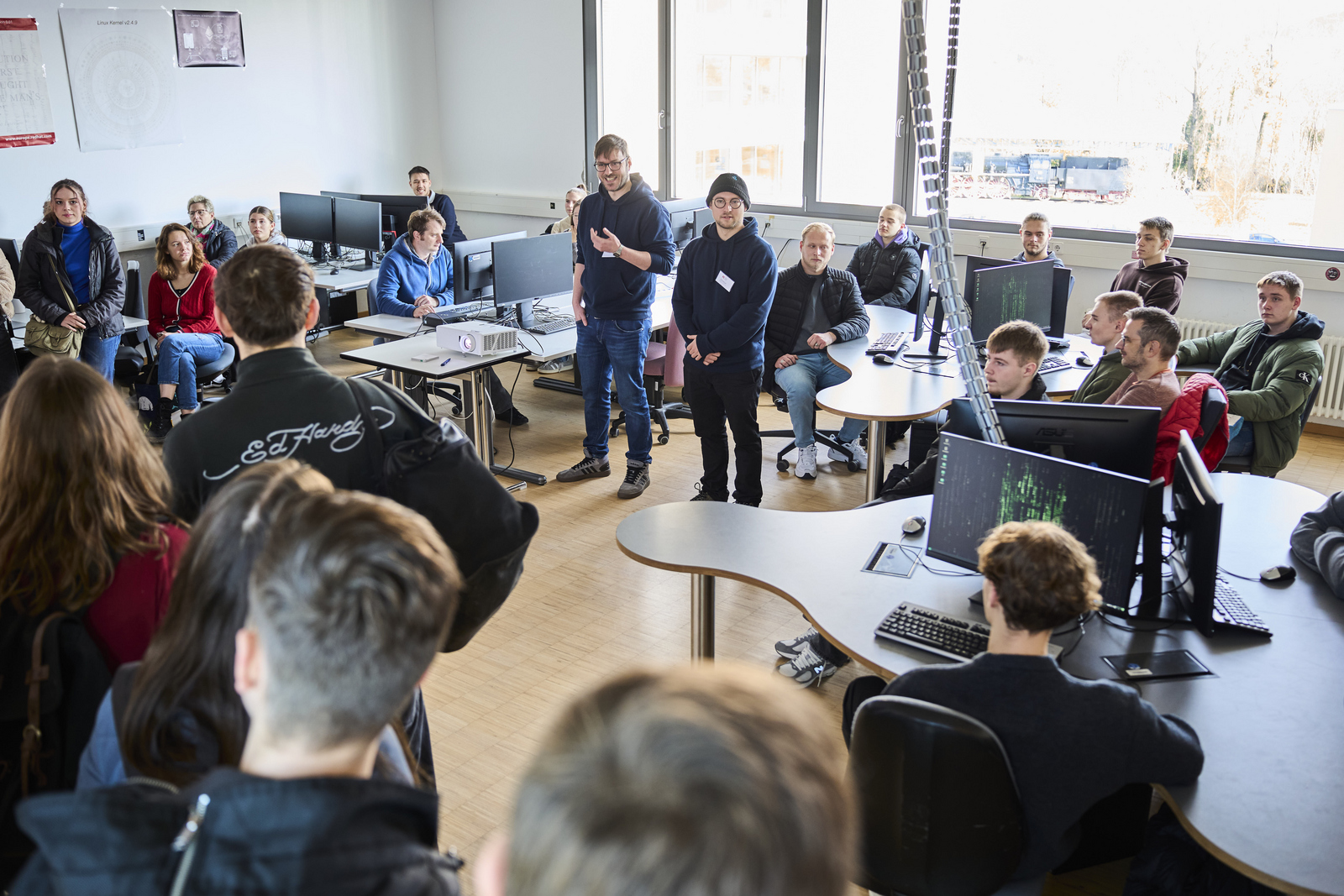 Zwei Studenten stehen umringt von sitzenden und stehenden Schülerinnen und Schülern in einem Computerraum und erzählen etwas