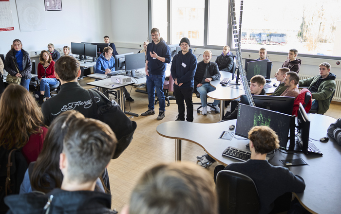 Zwei Studenten stehen umringt von sitzenden und stehenden Schülerinnen und Schülern in einem Computerraum und erzählen etwas